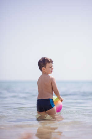 Adorable boy standing in the water on the beach holding a colorful beach ball in handsの写真素材