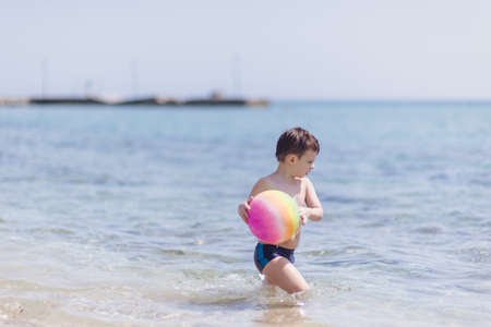 Adorable boy standing in the water on the beach playing with colorful ball and having funの写真素材