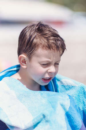 Wet young boy sitting on the beach with towel around his neck getting some heat in the sunの写真素材