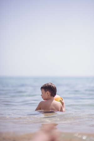 Adorable boy standing in the water on the beach holding a colorful beach ball in handsの写真素材