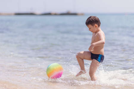 Adorable boy standing in the water on the beach playing with colorful ball and having funの写真素材