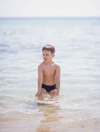 Adorable boy that wears black swimsuit is standing in the shallow water on the beachの写真素材