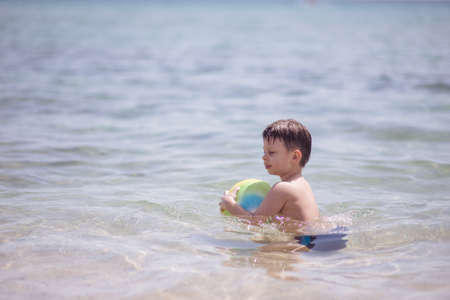 Adorable boy standing in the water on the beach holding a colorful beach ball in handsの写真素材