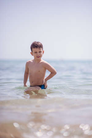 Adorable boy that wears black swimsuit is standing in the shallow water on the beachの写真素材