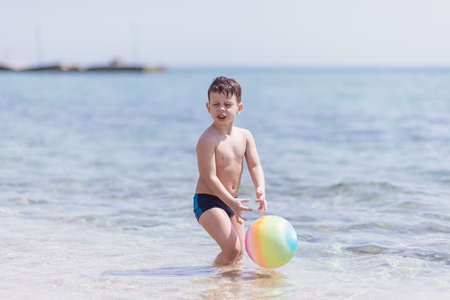 Adorable boy standing in the water on the beach playing with colorful ball and having funの写真素材