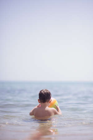 Adorable boy standing in the water on the beach holding a colorful beach ball in hands