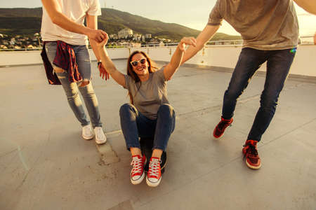 Three carefree friends playing with longboard on the balcony of the building at sunset