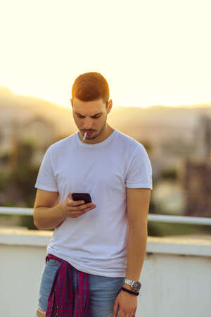 Young man holding his cellphone while smoke cigarette on the balcony in summer timeの写真素材