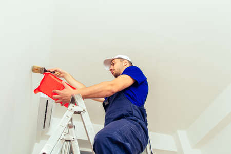 Young male decorator with paintbrush standing on scale while coloring wall