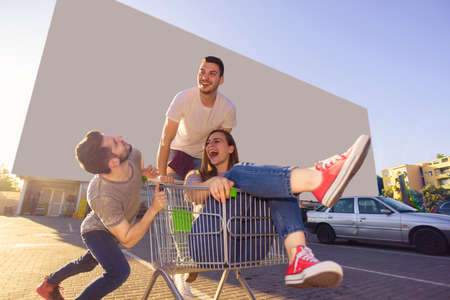 Two guys driving shopping trolley with girl in it on a parking lot