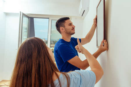 Positive man and woman hanging art picture in frame in their new living roomの写真素材