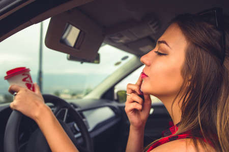 Pretty young woman is in her car holding coffee and fixing her lipstickの写真素材