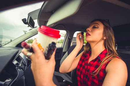 Girl is holding her makeup and coffee in her hands and enjoy applying make up on the front seat of...
