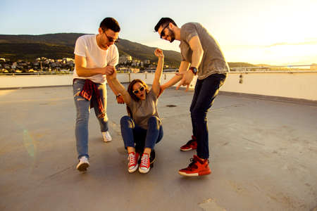 Three carefree friends playing with longboard on the balcony of the building at sunsetの写真素材