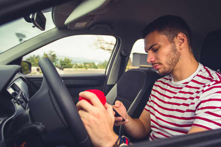 A guy is sitting in his car while holding coffee in one hand and looking interested in his mobile phoneの写真素材