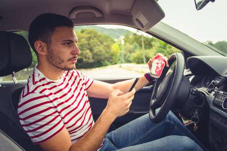 Young man is looking formard while is sitting in his black car and he is having coffee in one hand and his phone in the otherの写真素材