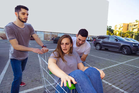 Cheerful friends amused on parking lot while driving a girl who sitting in a shopping cartの写真素材