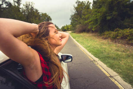A girl is with her head out of a car window while is driving in nature, smiling and having funの写真素材