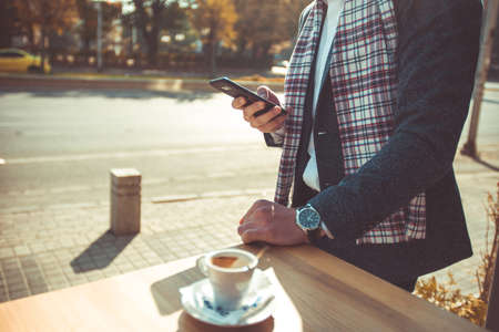 A stylish guy is standing near the table in the cafe, looking at his phone and having a coffeeの写真素材