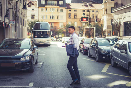 Young businessman is walking across the street in an urban city, holding his coffee and notebook and enjoying the beautiful weatherの写真素材