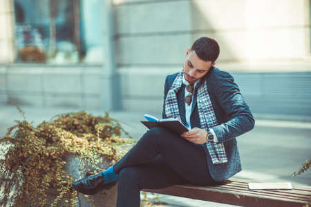 A stylish man is sitting with crossed legs on the bench, talking on his phone and writing in the notebookの写真素材