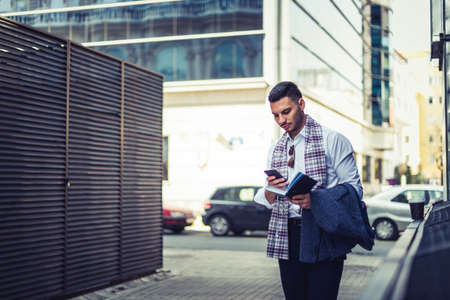 Confident male businessman is standing on the street and looking curious in his phone with opened notebookの写真素材