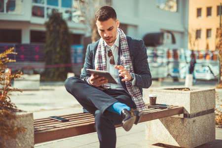 A good looking business man is enjoying the beautiful weather while sitting on the city bench with his coffee and tabletの写真素材