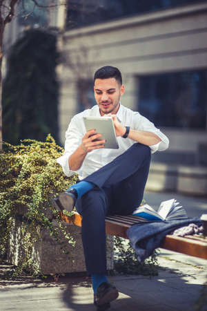 Urban businessman is sitting on the bench in the city center, looking at his tablet and enjoying the good weatherの写真素材