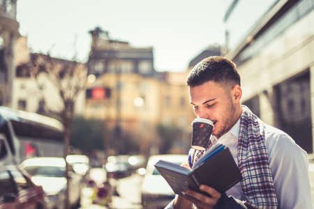 An urban businessman is being outdoors on a sunny day, drinking his coffee and writing in his notebookの写真素材