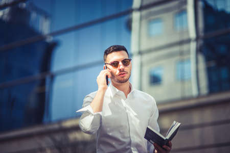 Urban businessman is making a phone call while walking near buildings with glasses, with sunglasses on and notebook in his handsの写真素材