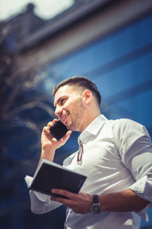 A young businessman is standing on the city square enjoying the sun and having a phone conversation while looking at his notebookの写真素材