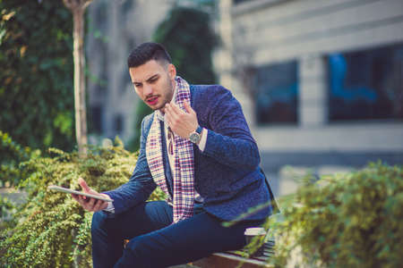 A man is sitting outside on the city bench and looking very serious at his tablet with his coffee near himの写真素材