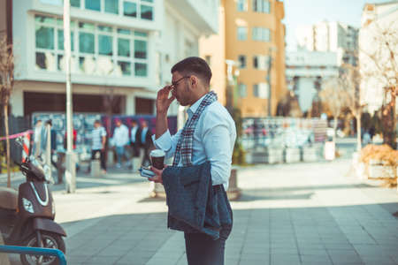 Urban businessman in suit is standing on the city square, holding notebook and coffee and looking around himの写真素材