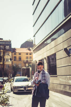 Urban businessman is standing in a beautiful city while drinking coffee and looking at his notebook