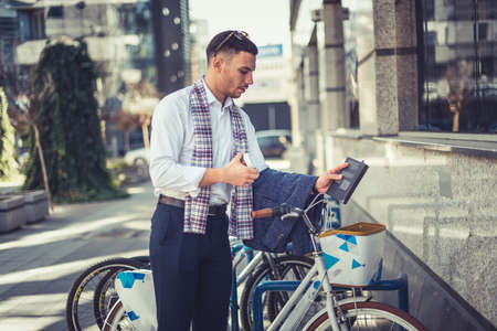 An urban man in business suit is leaning to a bike, holding a notebook and coffee in his handsの写真素材
