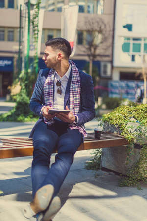 A man is sitting on the bench in the city center with crossed legs, holding tablet in his hands and looking aroundの写真素材