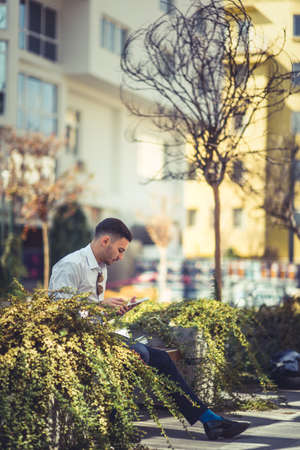 A stylish handsome man is sitting on bench in the city center and looking interested at his tabletの写真素材