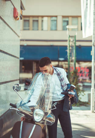 A handsome young businessman is standing near the motorbike, wearing a grey coat and having notebook and coffee in his handsの写真素材