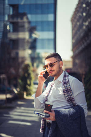 Urban businessman is having a phone call and looking satisfied while holding notebook and coffee in his handsの写真素材