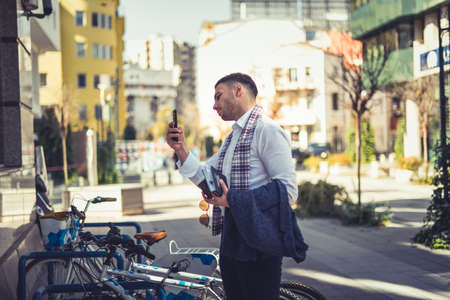 An urban businessman standing on the square next to his bike and taking picture on the glass mirror of the buildingsの写真素材
