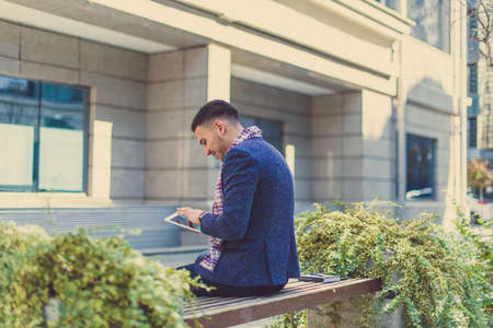 A man is sitting in front of some buildings, looking satisfied and happy at his tablet