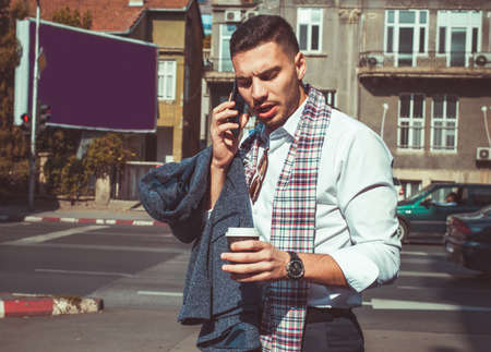 Confident lawyer is walking on the street in the middle of the city jam and having a phone call, while holding coffee and coat in his handsの写真素材