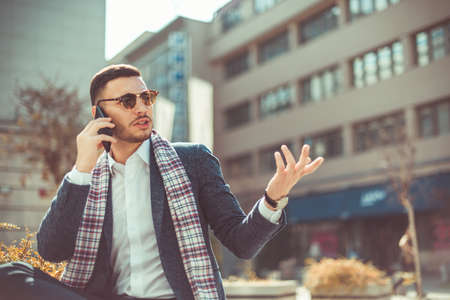 An urban businessman is having a conversation on his phone and arguing with his hands up while sitting in the city center near beautiful buildingsの写真素材