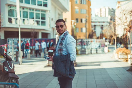 Urban businessman in suit is standing on the city square, holding notebook and coffee and looking around himの写真素材