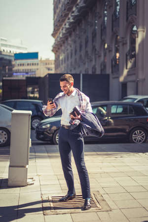 An urban man is in public, standing on the street and looking at his phone on a windy dayの写真素材