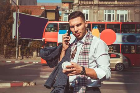 Handsome businessman is walking in the middle of the street, holding a coffee and phone in his hands in the middle of city jamの写真素材