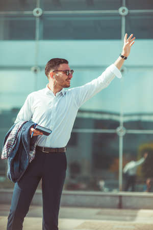 Cheerful young businessman is standing in urban city, waving with his hand up on someone and smilingの写真素材