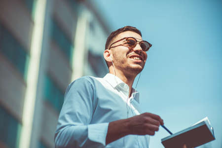 A confident businessman is in urban neighbourhood with his headphones on, holding a pen and holding his notebookの写真素材