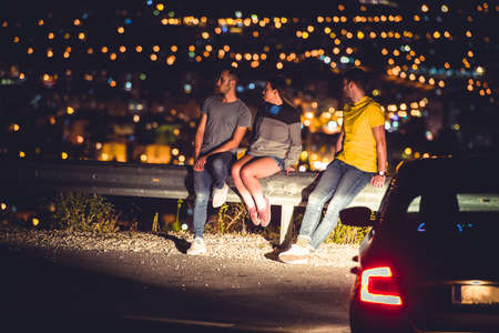 Friends are sitting on the road guardrail at night enjoying the city panorama and lightsの写真素材