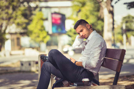 A modern businessman is sitting in the park on a bench with crossed legs and he is looking curious and seriousの写真素材